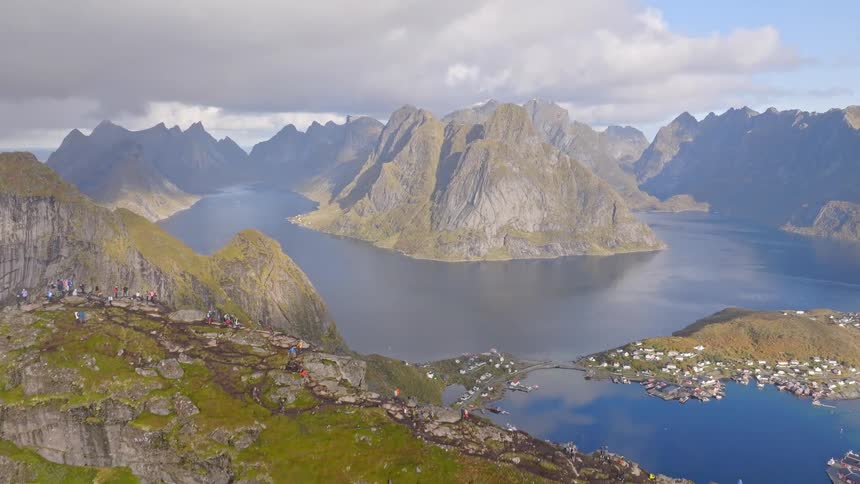 Epic aerial pullback reveals hikers on a mountain summit enjoying the breathtaking view of Norwegian fjords and a scenic fishing village