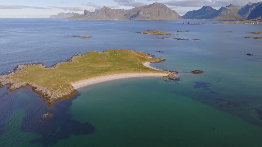 Aerial view tilts up to reveal a stunning mountain coastline behind a secluded, grassy island with a beautiful sandy beach