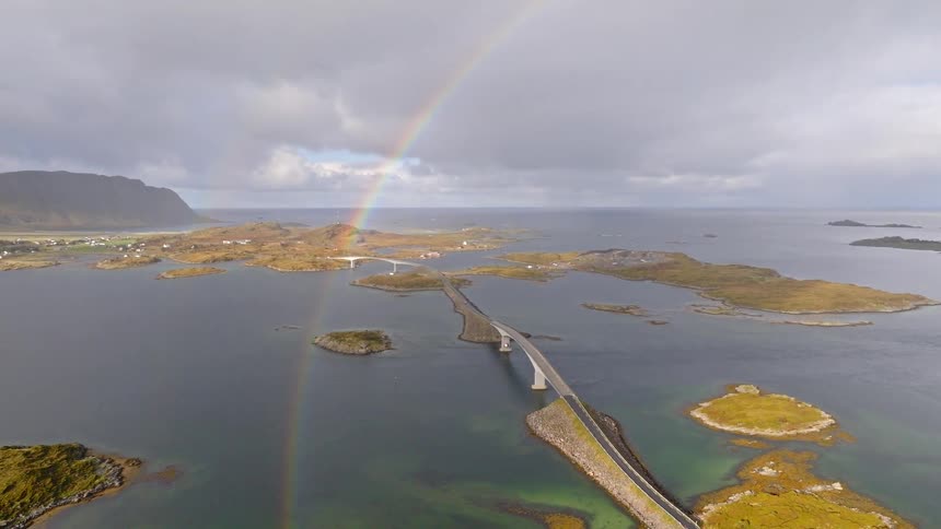 A slow aerial pan reveals a magnificent double rainbow over a long bridge connecting a series of remote islands in Norway