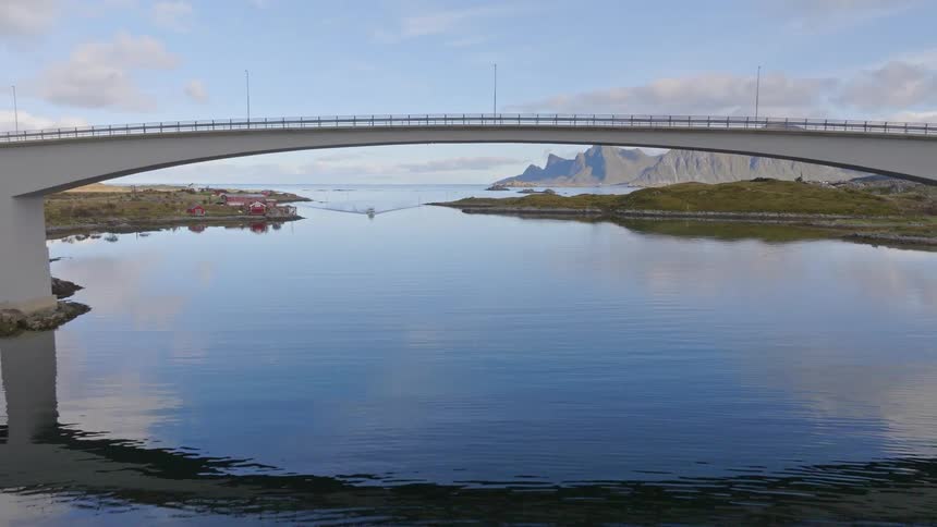 A stunning aerial reveal shot flying backward under a modern bridge over a calm Norwegian fjord with mountains in the background