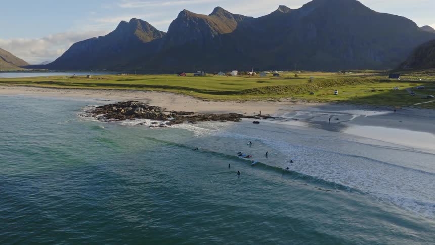 Stunning aerial pan of surfers waiting for waves on a scenic beach at the base of majestic mountains during a beautiful sunset
