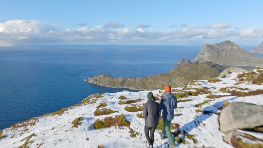 An aerial shot follows a couple as they hike along a snowy mountain ridge, revealing a breathtaking view of the Norwegian fjords and the vast ocean