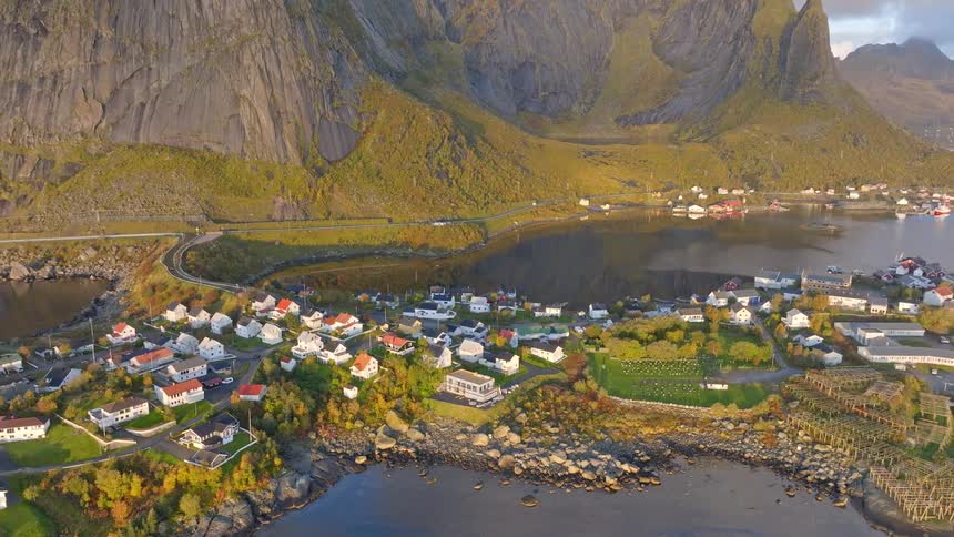 Aerial footage slowly pulls back to reveal a stunning Norwegian fishing village nestled between the sea and colossal mountains during a golden autumn sunrise