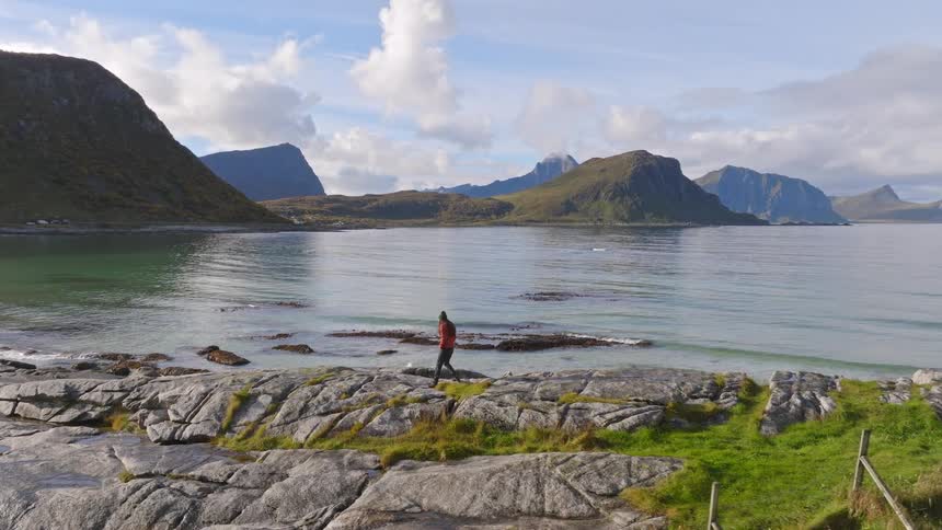 A person enjoys a scenic walk along a rocky shoreline next to a tranquil bay surrounded by towering mountains