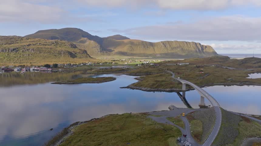 A majestic aerial shot showcases a winding bridge over calm waters, leading to a village nestled at the base of large mountains