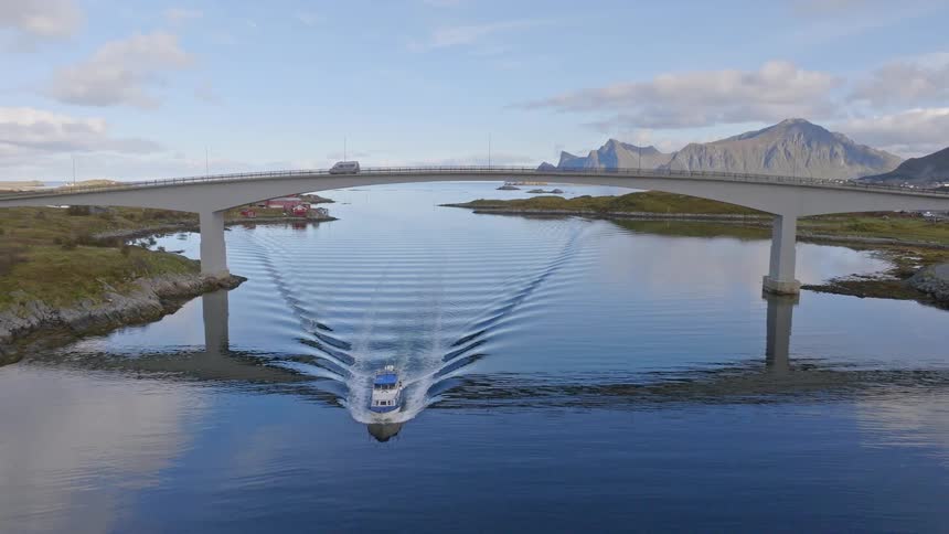 An aerial drone flies backward under a scenic bridge in Norway as a boat sails on the water below and a camper van crosses above
