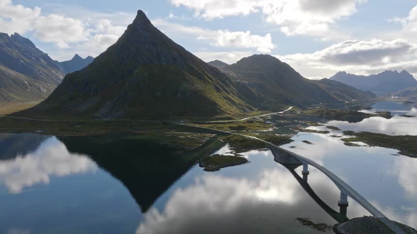 A majestic, pointed mountain and its reflection stand over a serene lake, with a modern bridge curving across the tranquil water