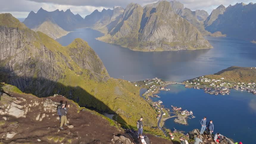 An incredible aerial shot pulls back to reveal a group of hikers on a mountain summit overlooking a beautiful fjord and village in Norway