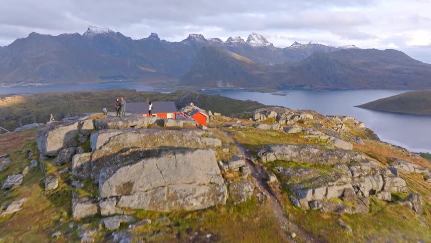 An aerial drone flies towards hikers on a rocky summit, revealing a remote red cabin overlooking a stunning Norwegian fjord and snow-capped mountains