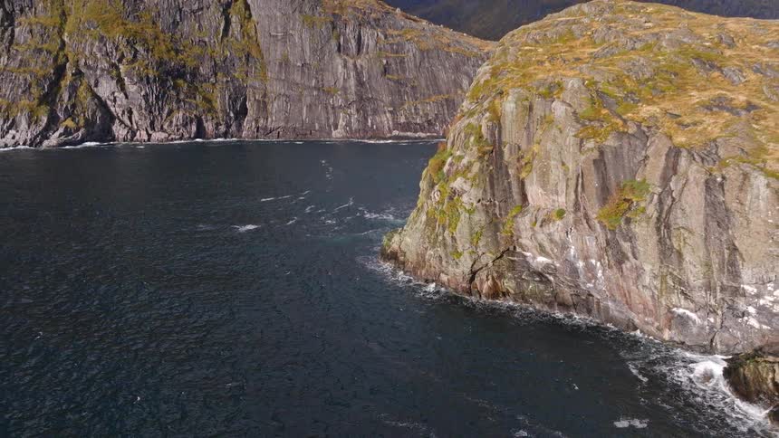 A slow pull-back shot reveals the dramatic, sheer cliffs and snow-dusted mountains surrounding a rugged fjord