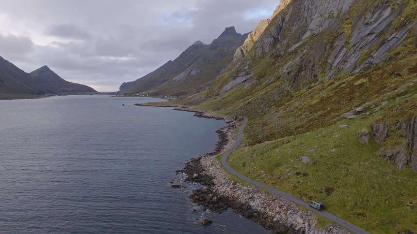 Aerial footage follows a camper van driving along a dramatic coastal road nestled between the sea and steep mountains
