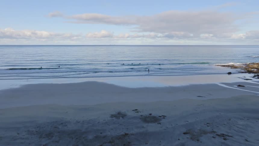 Drone flies past a modern coastal building and over grassy dunes to reveal surfers riding gentle waves in a scenic bay surrounded by mountains