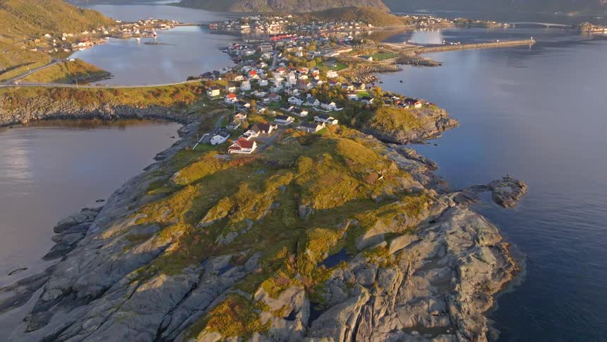 An aerial drone shot tilts up to reveal the picturesque fishing village of Reine in the Lofoten Islands, Norway, nestled among dramatic mountains and fjords at sunrise