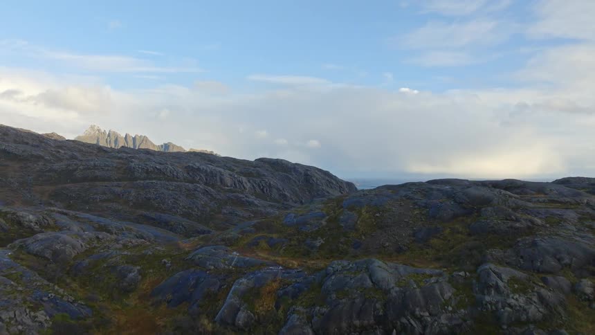A drone slowly ascends over a vast, rocky Norwegian landscape, revealing pristine blue lakes, the ocean, and a dramatic mountain range on the horizon