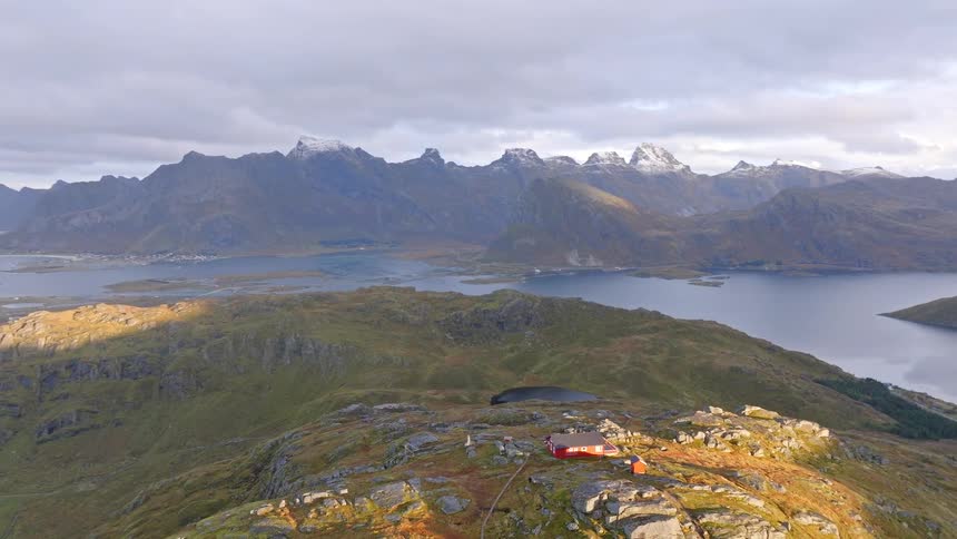 Epic aerial view slowly rises over a vast Norwegian fjord landscape with a lone red cabin perched on a sunlit hill