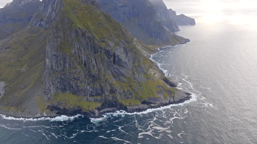 A stunning aerial tilt-up reveals a rugged, moss-covered mountain range meeting the restless ocean along a dramatic coastline