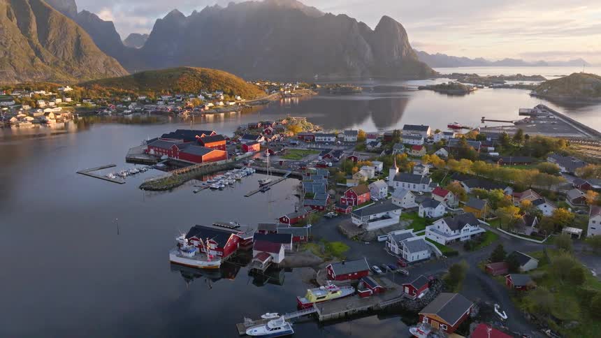 An aerial view slowly ascends over a quaint fishing village in Lofoten, Norway, showcasing the beautiful harbor against a backdrop of majestic mountains at sunrise