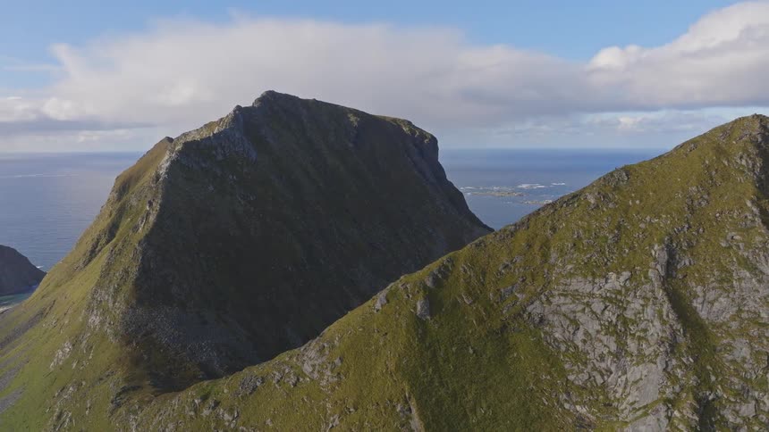 Aerial view rises over a dramatic green mountain ridge to reveal a pristine lake nestled between peaks overlooking the vast ocean