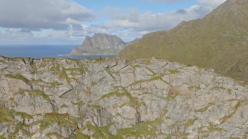 A breathtaking aerial orbit reveals hikers standing atop a dramatic mountain ridge overlooking a tranquil lake and the vast ocean in the Lofoten Islands, Norway