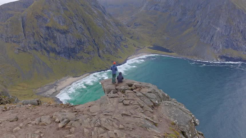 Two adventurous hikers stand on a dramatic cliff edge, overlooking a stunning turquoise ocean bay and mountainous coastline