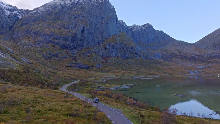 An aerial view shows a campervan driving on a winding road alongside a lake, surrounded by towering, snow-dusted mountains in Norway