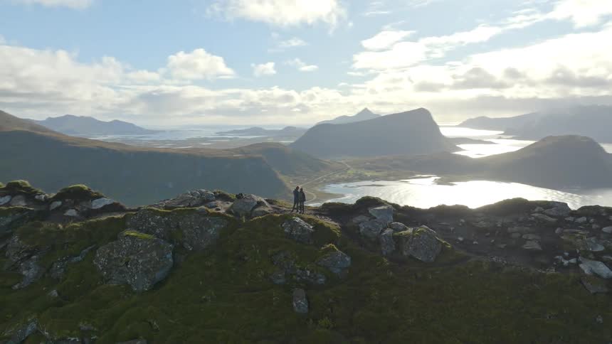 An aerial shot orbits a couple standing on a majestic mountain peak, revealing a stunning panoramic landscape of fjords and the vast ocean