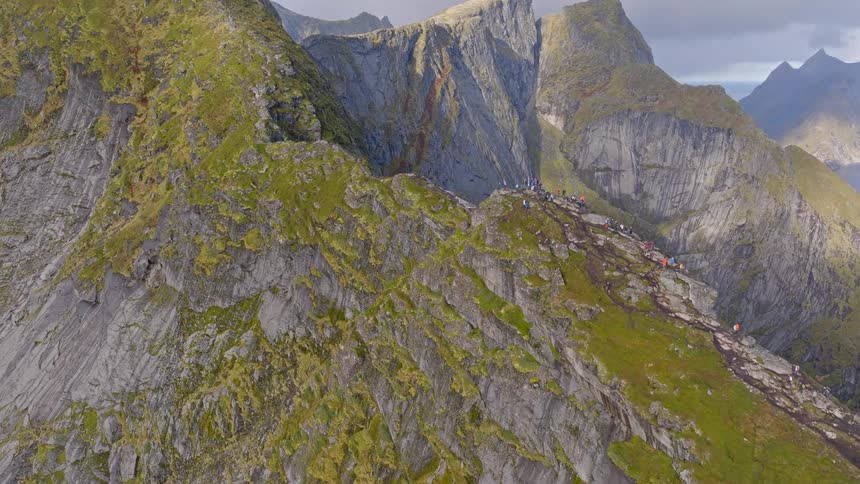 An epic aerial ascent reveals a crowd of hikers on the famous Reinebringen trail, showcasing the dramatic, steep ridges of Lofoten, Norway