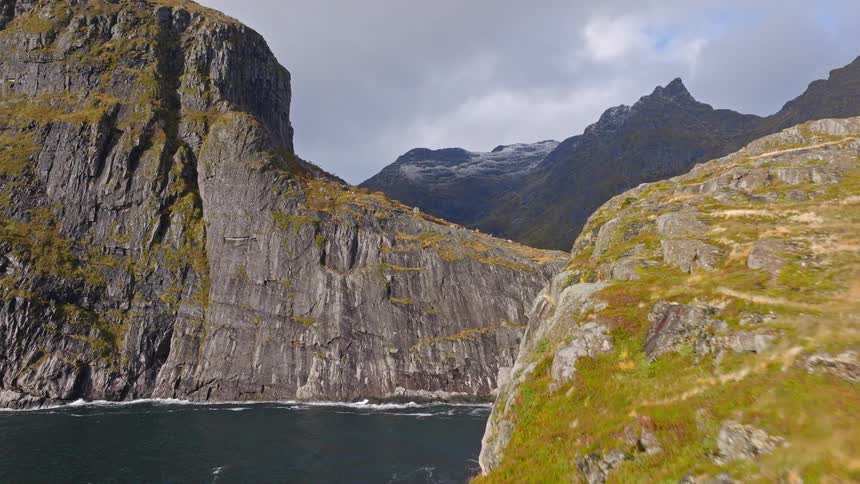 Epic aerial footage flies over a rugged, rocky cliffside revealing a stunning fjord and snow-capped mountains