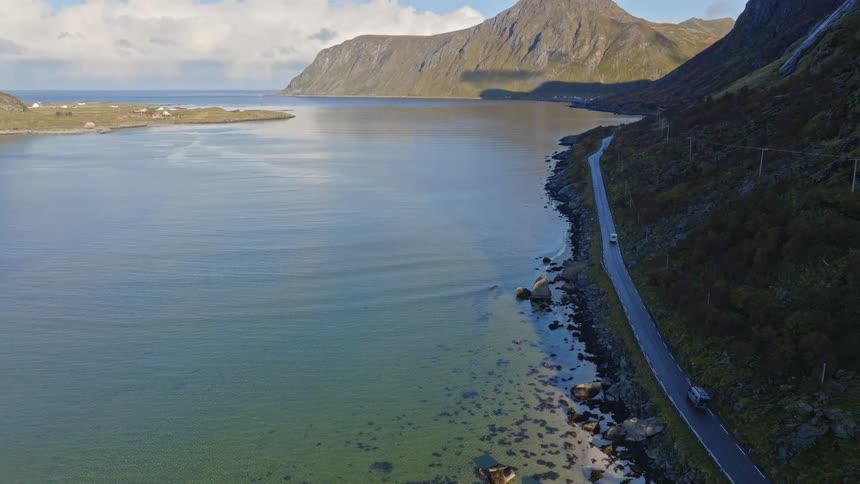 An aerial view shows vehicles driving on a scenic coastal road alongside a tranquil bay with majestic mountains in the background