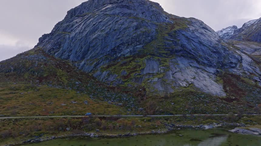 A blue camper van takes a scenic drive along a lakeside road at the foot of a towering, mossy mountain in Norway