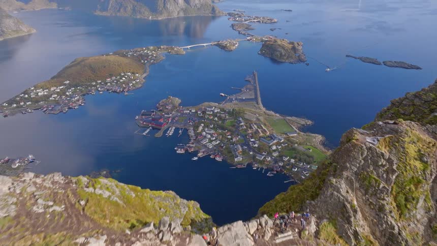 A stunning aerial view flies over the steep green ridge of Reinebringen mountain, revealing the picturesque fishing village of Reine in the Lofoten Islands, Norway