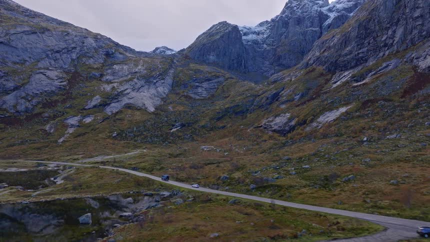 An expansive aerial shot captures cars navigating a winding road through a majestic mountain valley with snow-capped peaks