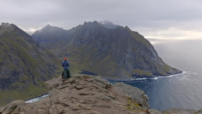 An aerial view reveals two hikers on a rocky mountain summit enjoying the breathtaking scenery of a secluded beach and the vast ocean