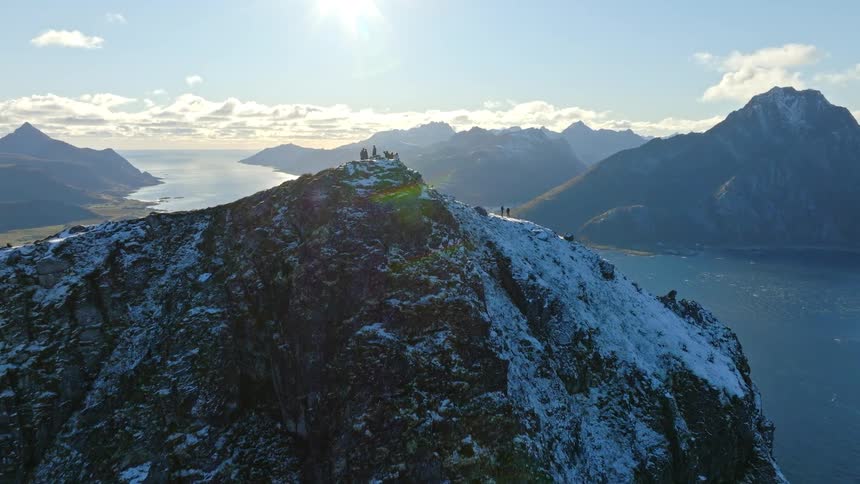An inspiring aerial view reveals hikers on a snowy mountain summit overlooking a vast, sunlit fjord