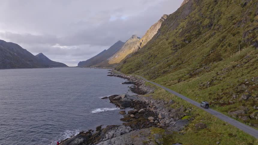 An aerial view shows a camper van driving on a scenic coastal road through the majestic fjords of Norway
