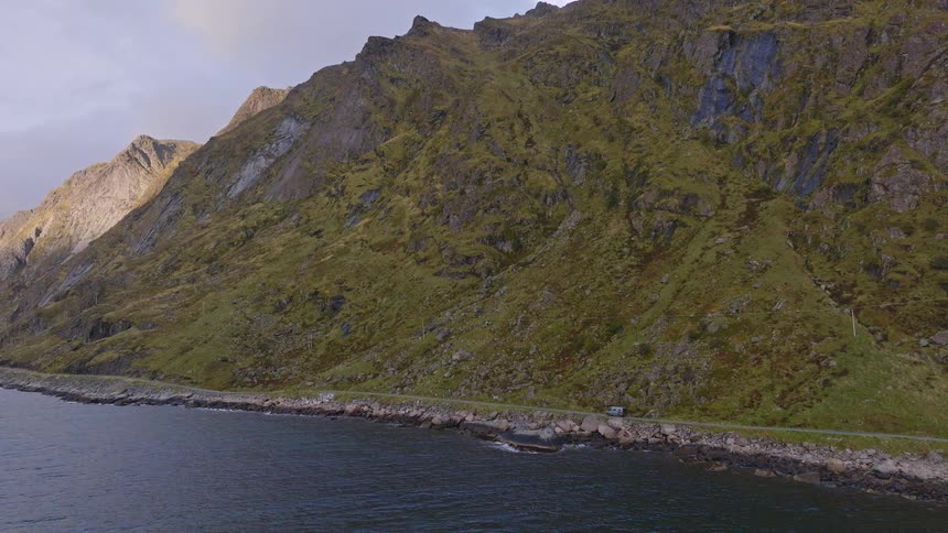 A static aerial view shows a camper van driving along a remote coastal road at the foot of a giant, moss-covered mountain in Norway