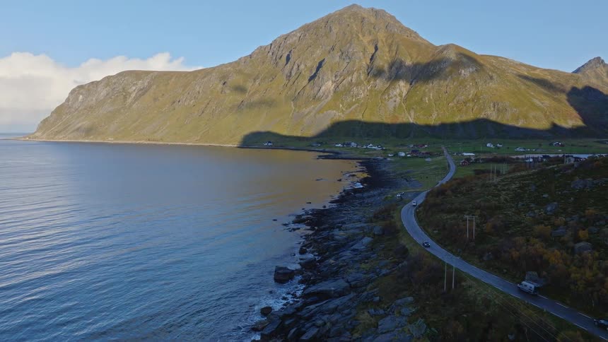 Aerial view of a scenic coastal road in Norway where cars drive alongside the ocean at the foot of a massive mountain on a sunny day
