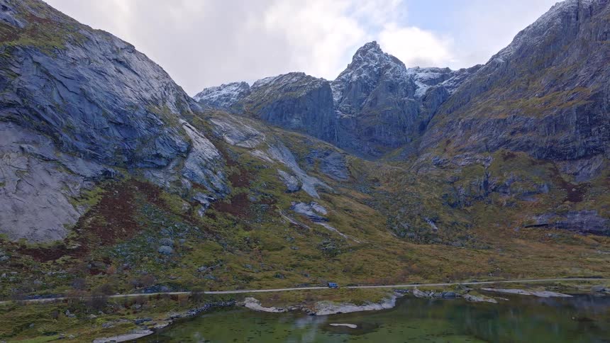 An epic aerial view of a lone car driving on a scenic road through a massive, snow-capped mountain valley next to a calm lake
