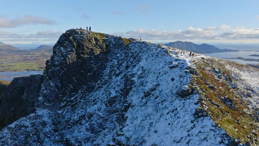 A stunning orbiting aerial shot reveals hikers on a snow-dusted mountain peak overlooking the majestic fjords and islands of Norway