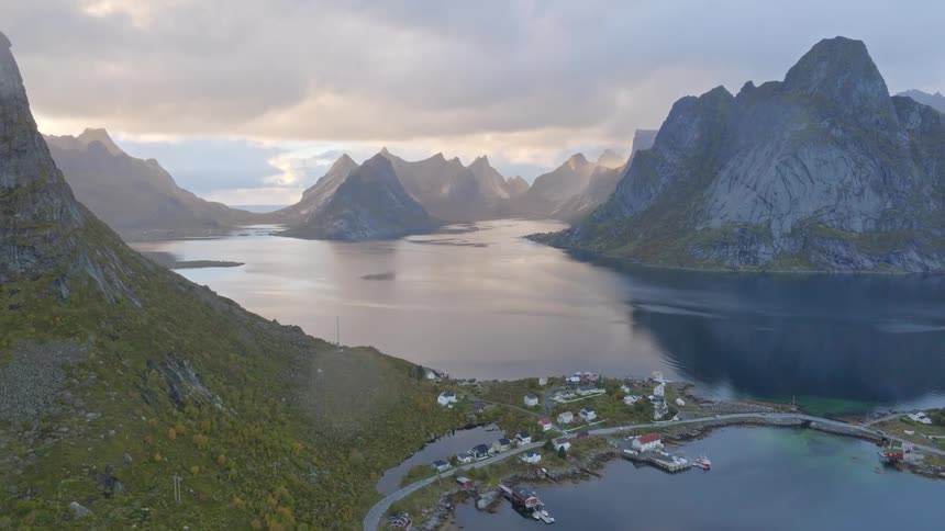 A dramatic aerial view pulls back to reveal a tranquil fishing village nestled within a stunning fjord, surrounded by majestic mountains in Norway