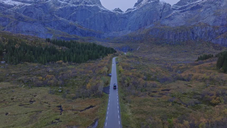 An aerial shot follows a camper van on a scenic road trip through a vast autumn valley towards majestic snow-capped mountains