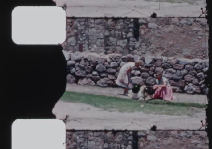 Vintage 16mm footage captures two women affectionately petting their dogs in a rustic stone courtyard on a sunny day