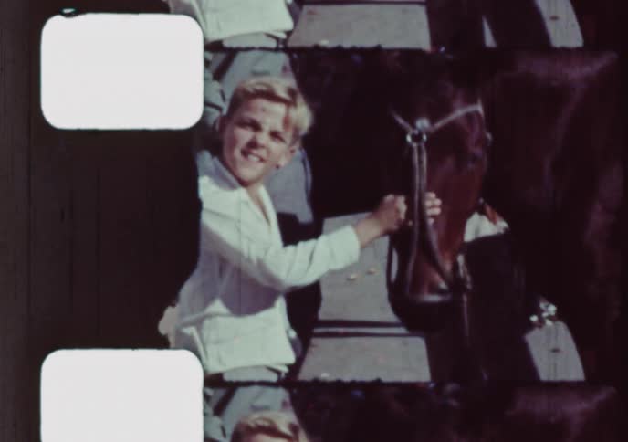 Vintage 16mm film captures a happy young boy with blond hair gently petting a calm horse on a sunny day