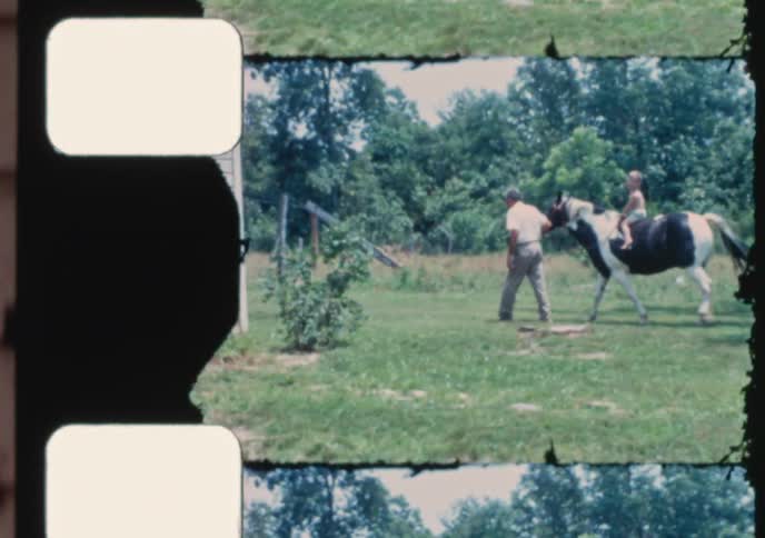 Vintage film footage shows a man leading a pinto horse with a young boy riding on its back through a grassy field