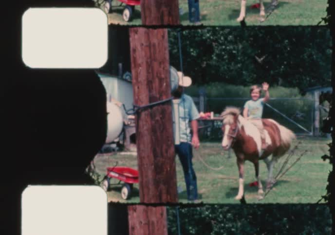 A father wearing a cowboy hat gives his smiling son a ride on a pony in a grassy backyard in this vintage home movie from the 1970s