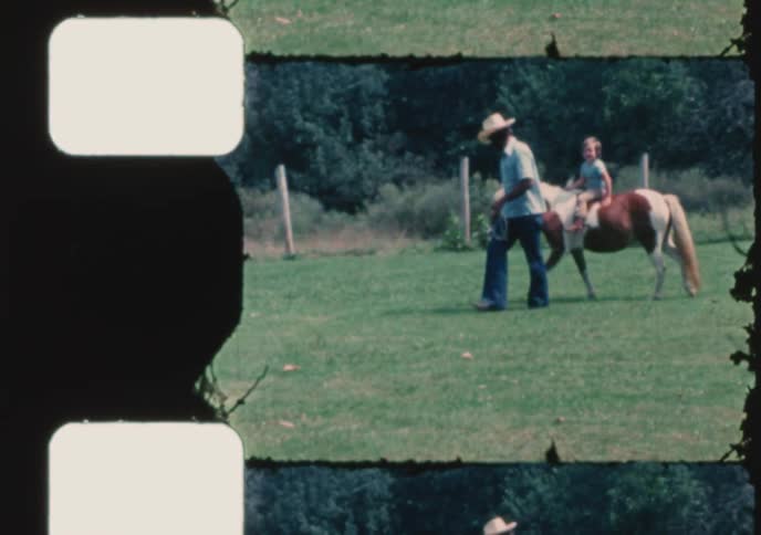 A man wearing a cowboy hat leads a small pony with a young child on its back through a grassy pasture in this vintage home movie clip