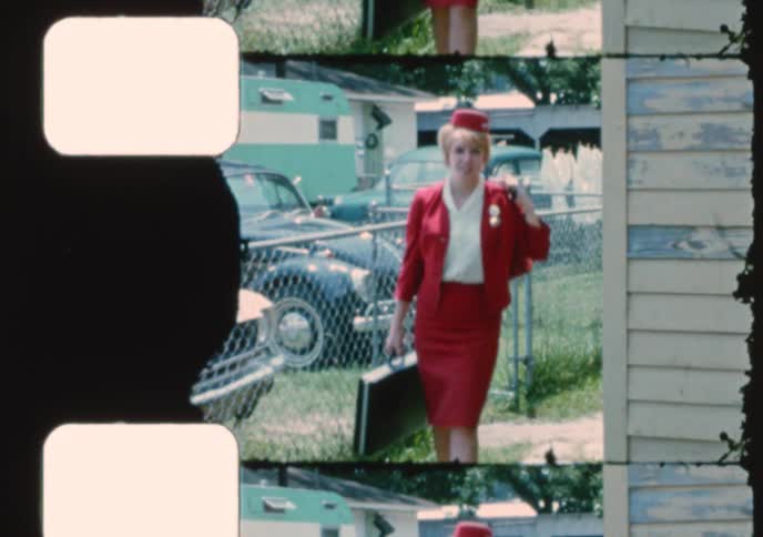 A happy flight attendant in a red uniform walks through a residential neighborhood in this 16mm film scan