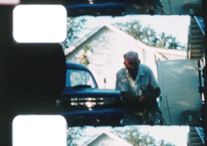 An elderly man playfully pretends to ride a shovel in front of his vintage truck in an old 8mm home movie