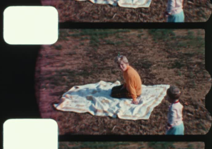 A mother sits on a picnic blanket in a field as her toddler son walks and crawls nearby in this nostalgic home movie clip