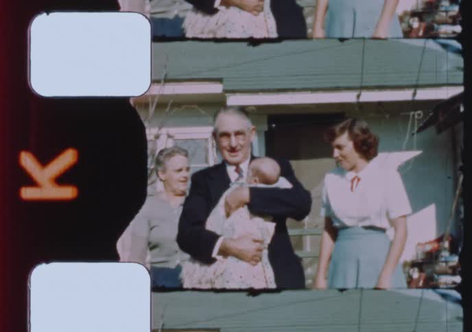 A proud grandfather holds his newborn grandchild while surrounded by family in a vintage home movie from the 1950s